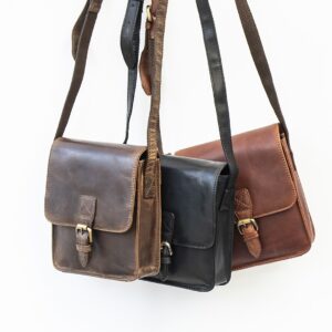 three leather man bags are hanging up. the bags have detailed stitching to the front with a brass buckle