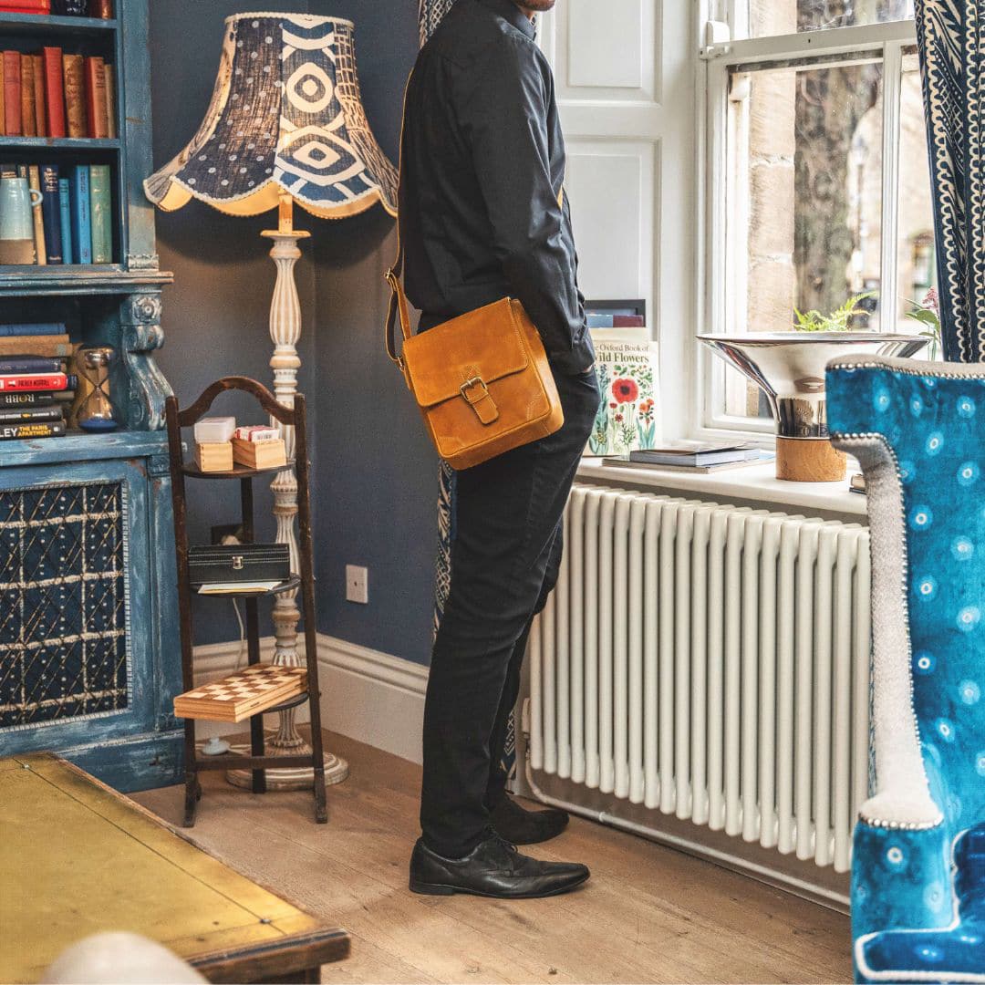 a man is wearing a mustard coloured leather messenger bag. the bag has a centre brass buckle