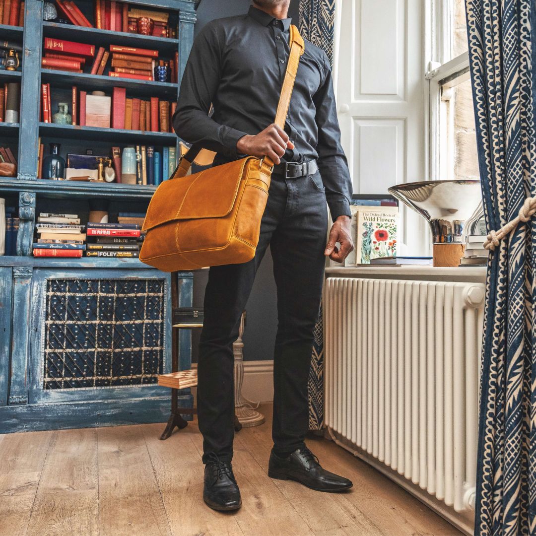 a man is wearing a luxury leather messenger bag is a crossbody style. The bag is mustard in colour with detailed stitching. the man is dressed in black with a black leather belt accessory