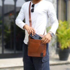 Man placing phone in front pocket of chestnut leather messenger bag – stylish shoulder bag for men and women, ideal for travel or work.