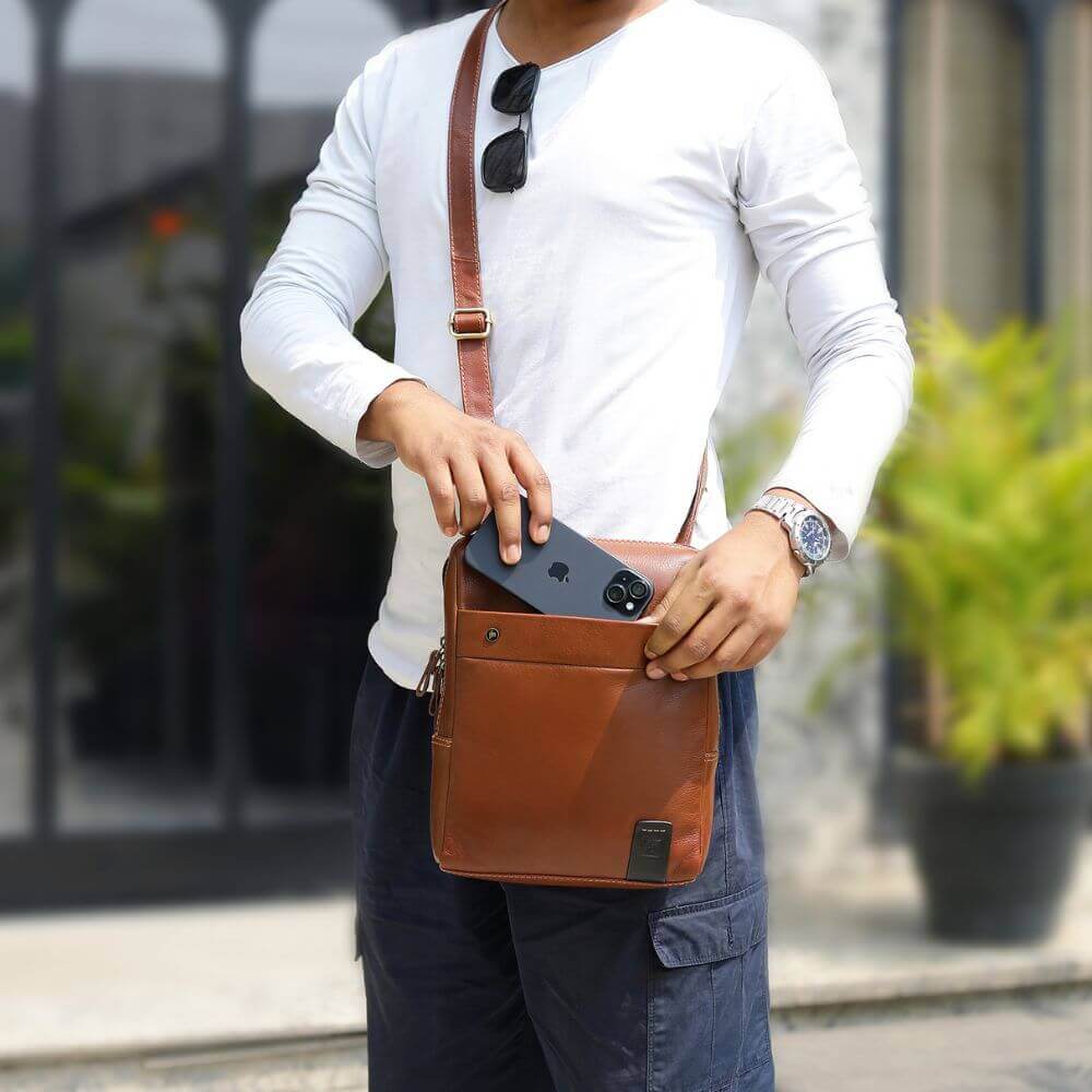 Man placing phone in front pocket of chestnut leather messenger bag – stylish shoulder bag for men and women, ideal for travel or work.
